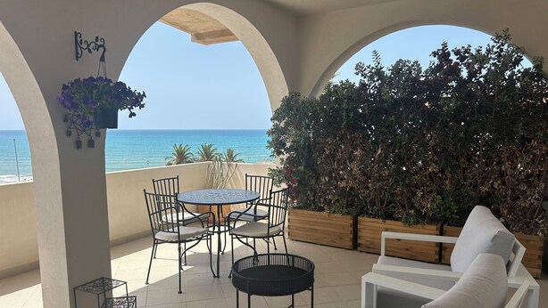 Un espace extérieur abrité, aménagé avec des sièges, offrant une vue sur la mer, une table ronde avec des chaises et un canapé blanc.
