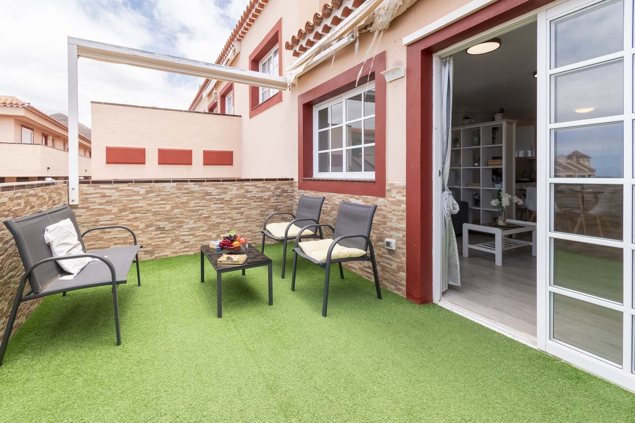 A balcony with artificial grass, two chairs, a small table with fruits, and a white pergola.