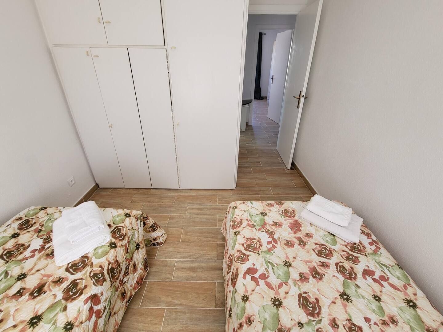 A small, clean room with two single beds, a wardrobe, and a hallway leading to another room.