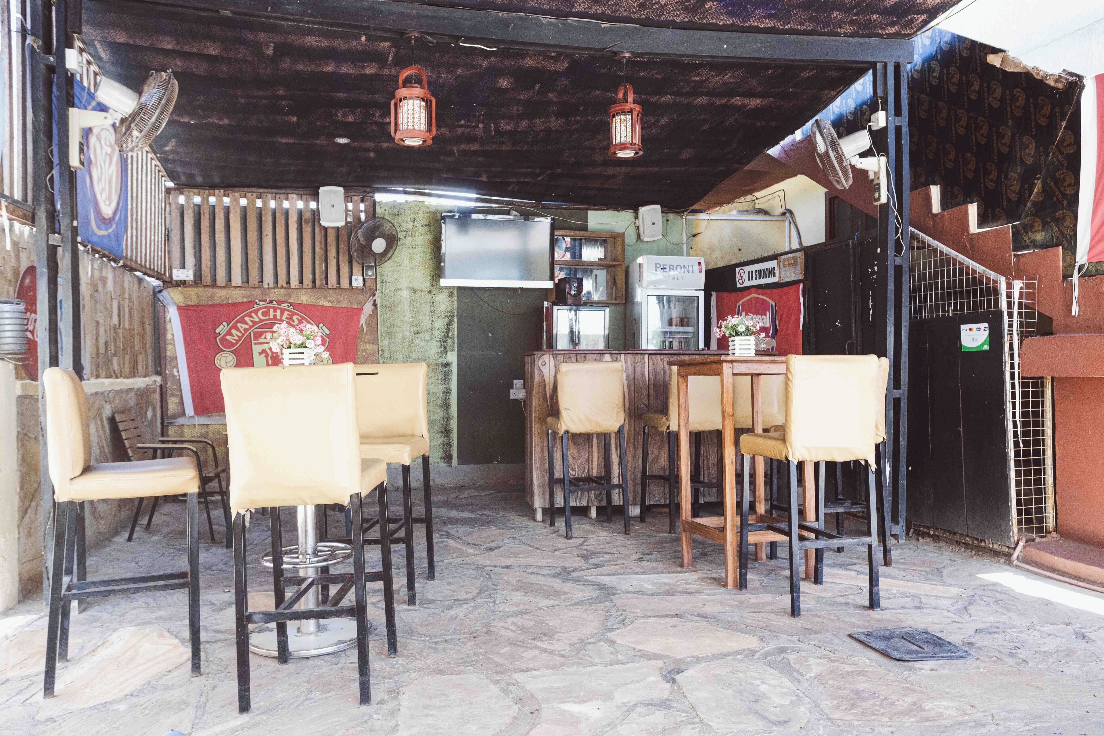 Un cafè a l'exterior amb sòl de pedra, taules i cadires de fusta, un refrigerador i un cartell amb la inscripció "Peroni" a la paret.
