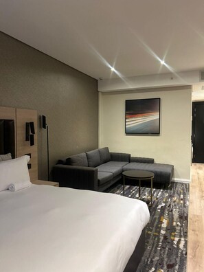 A modern hotel room with a large bed, a gray sectional sofa, a small round table, and a painting on the wall. - Luxury Apartment Sandton / no Loadshedding / Dstv (Johannesburg)