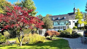 A traditional building with a prominent tower, surrounded by a well-maintained garden with various trees and shrubs. - Holiday Home Farmhouse in Altes Forsthaus Boppard (Boppard)