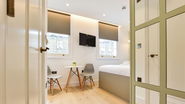 Ein kleines Zimmer mit einem Bett, einem an der Wand befestigten Fernseher, einem kleinen Tisch mit zwei Stühlen und einem Fenster mit Blick auf Gebäude.