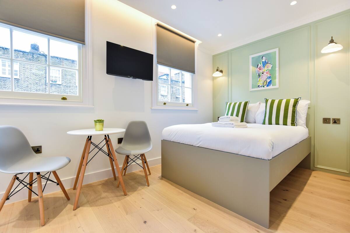 Un dormitorio moderno con una cama, dos sillas, una mesita, un televisor y una ventana con vistas a edificios.