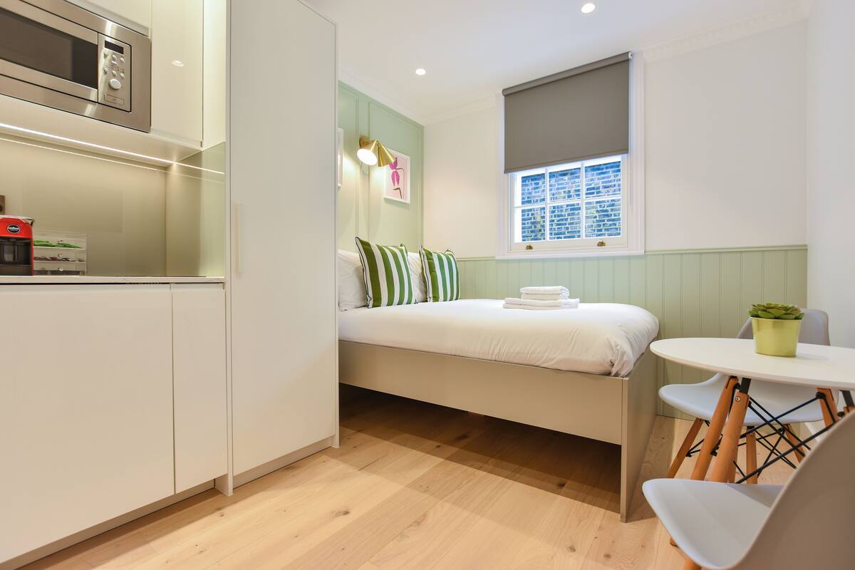 Una habitación compacta con cama, una mesita, una silla, microondas y ventana con persianas.