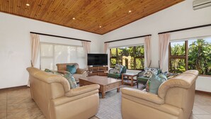 A spacious living room with leather sofas, a wooden coffee table, and a flat-screen TV. Large windows provide natural light and views of greenery. - San Lameer Villa 10438 (Southbroom)