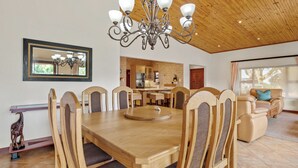 A dining area with a wooden table and chairs, a chandelier, a kitchen in the background, and a living area with a sofa and a window. - San Lameer Villa 10438 (Southbroom)