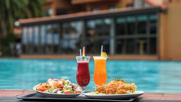 A dining table with a salad, a red drink, and a yellow drink by a poolside.