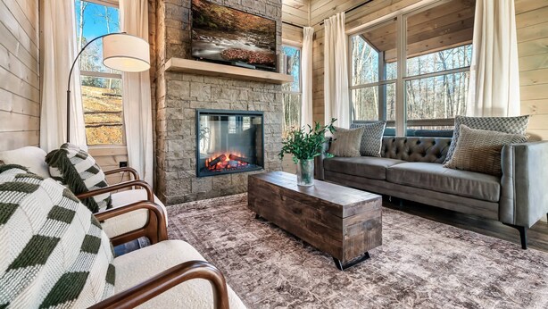 A cozy log cabin interior with a stone fireplace, a large TV mounted above it, a comfortable sofa, and a rustic wooden coffee table.