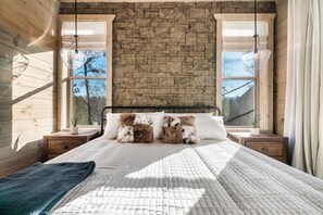 A bedroom with a stone wall, wooden floor, a bed with a quilt, two bedside tables, and a window with curtains. - Luxury Cabin w/ View・Pool・Hot Tub・Games・Fire pit (Gatlinburg)