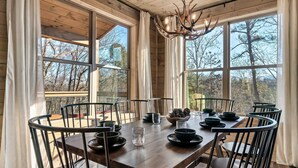 A wooden dining table with chairs, a chandelier, and large windows with curtains.