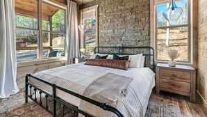 A bedroom with a large bed, a nightstand, a stone wall, and large windows.
