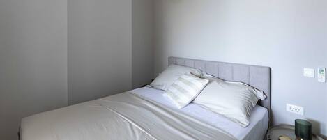 A neatly made bed with white linens and pillows, a grey headboard, and a bedside table with a green lamp.