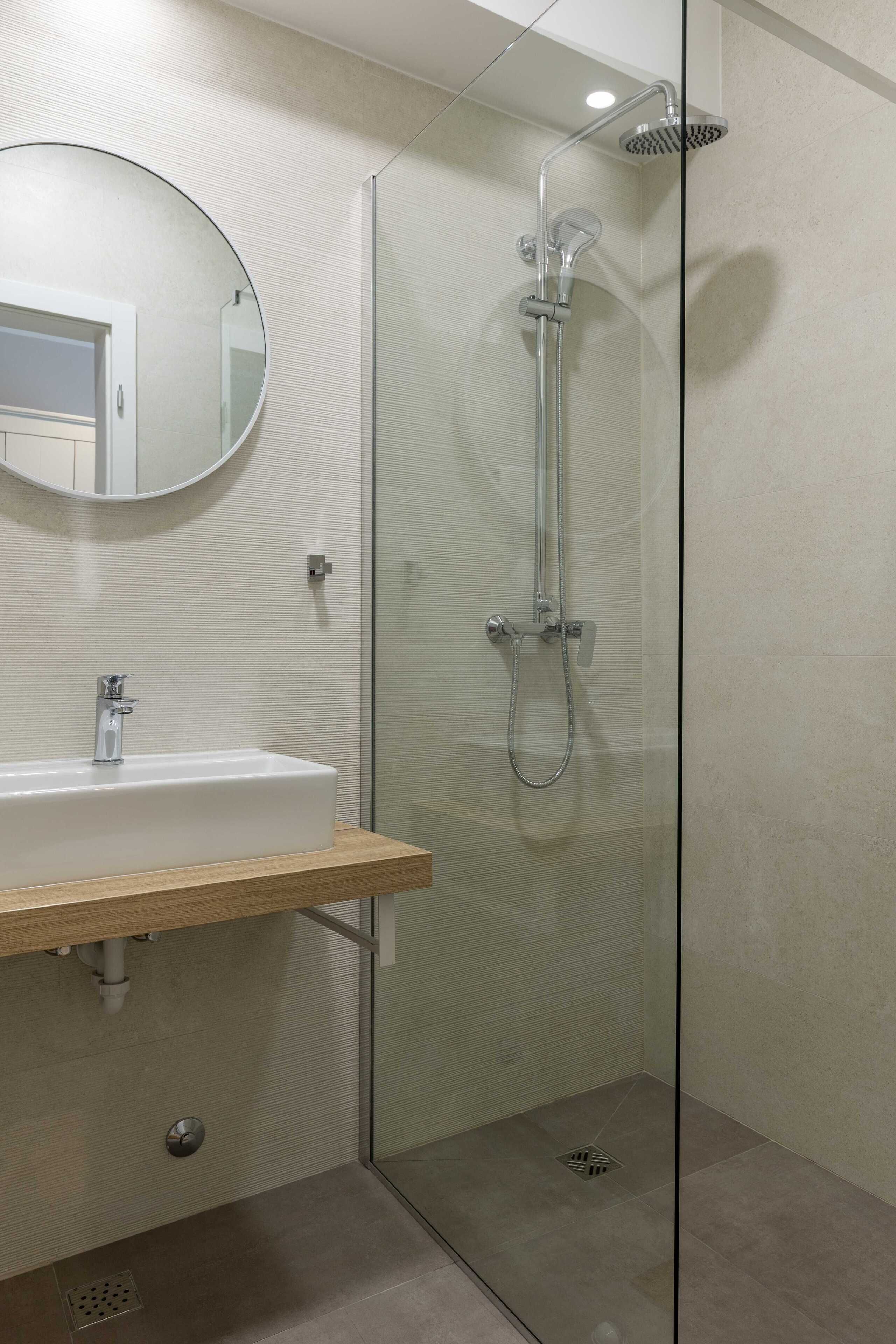 A modern bathroom with a glass-enclosed shower, a round mirror, and a wooden vanity.
