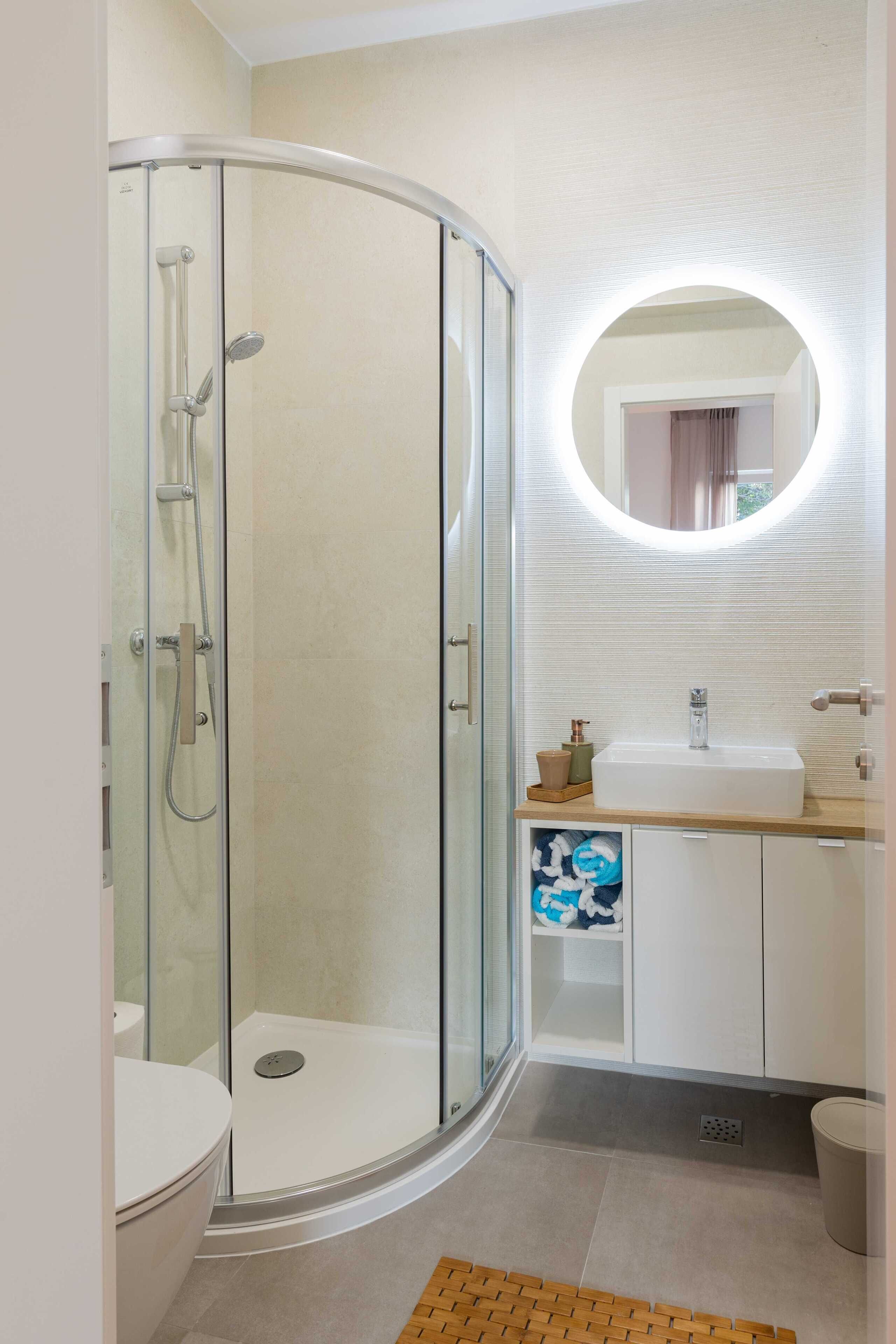 A modern bathroom with a glass-enclosed shower, a round mirror, and a white sink.
