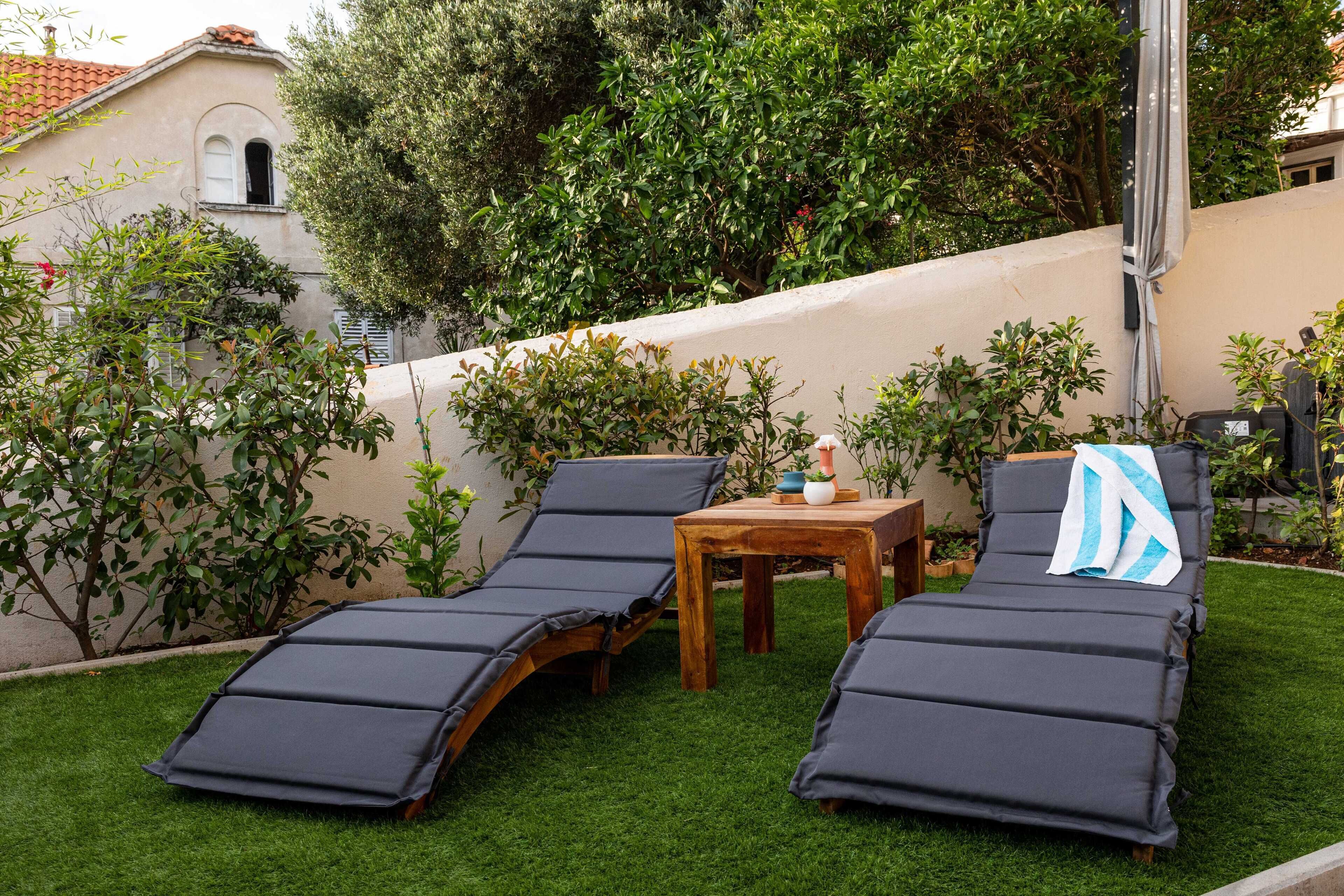 Two lounge chairs with cushions, a small wooden table, and a potted plant in a garden area with a white wall and greenery in the background.
