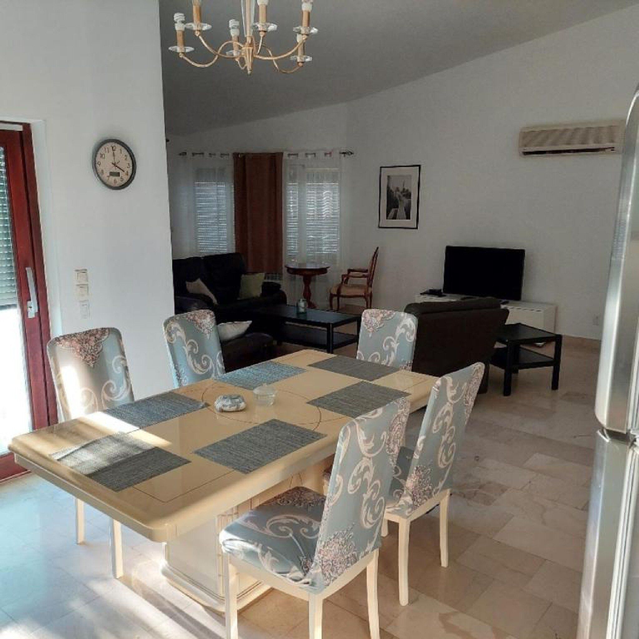 Un coin repas avec une table dressée pour quatre personnes, entourée de chaises à motifs. Un lustre est suspendu au plafond, et une horloge est visible au mur.