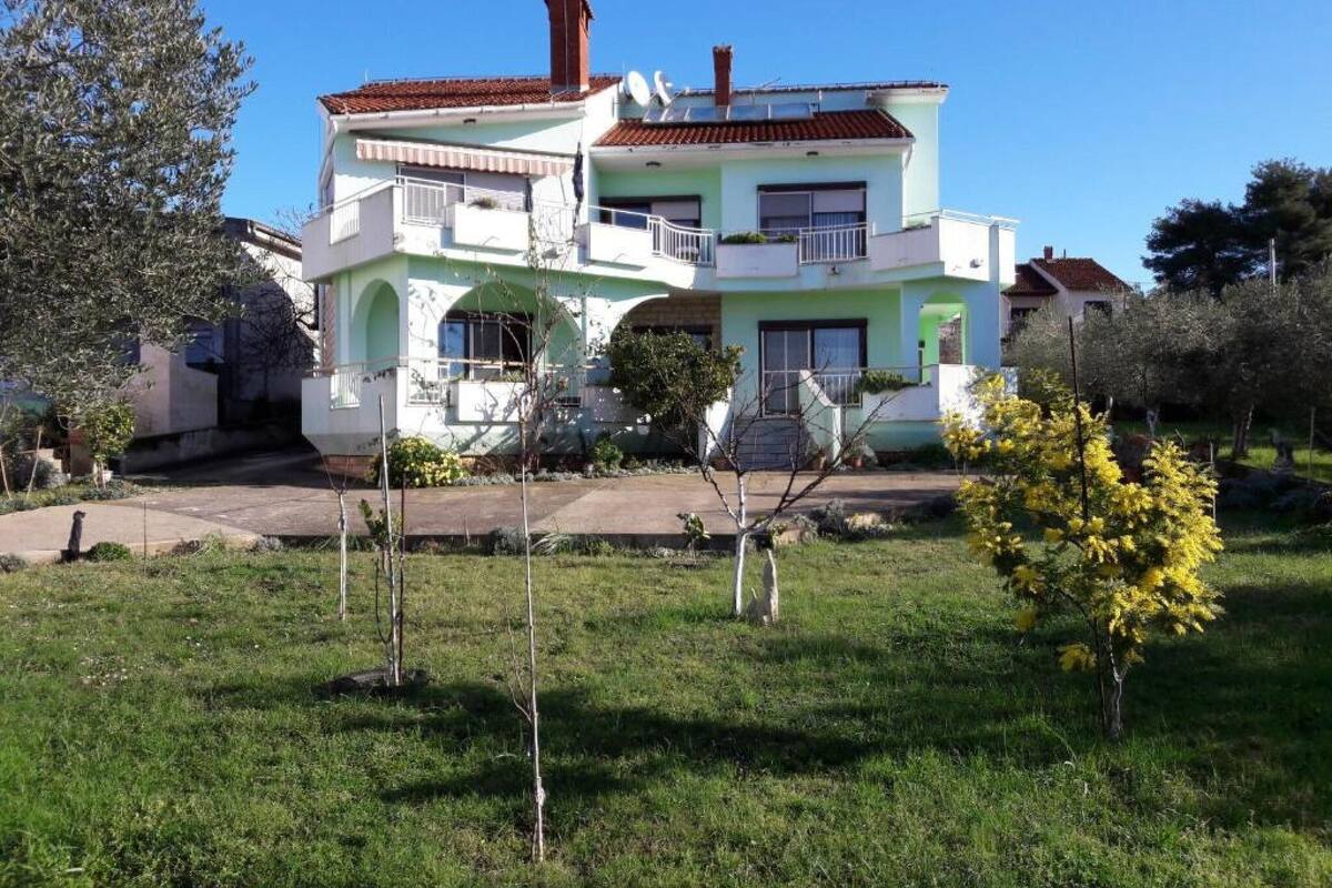 A two-story house with a balcony, a chimney, and a garden with yellow flowers.