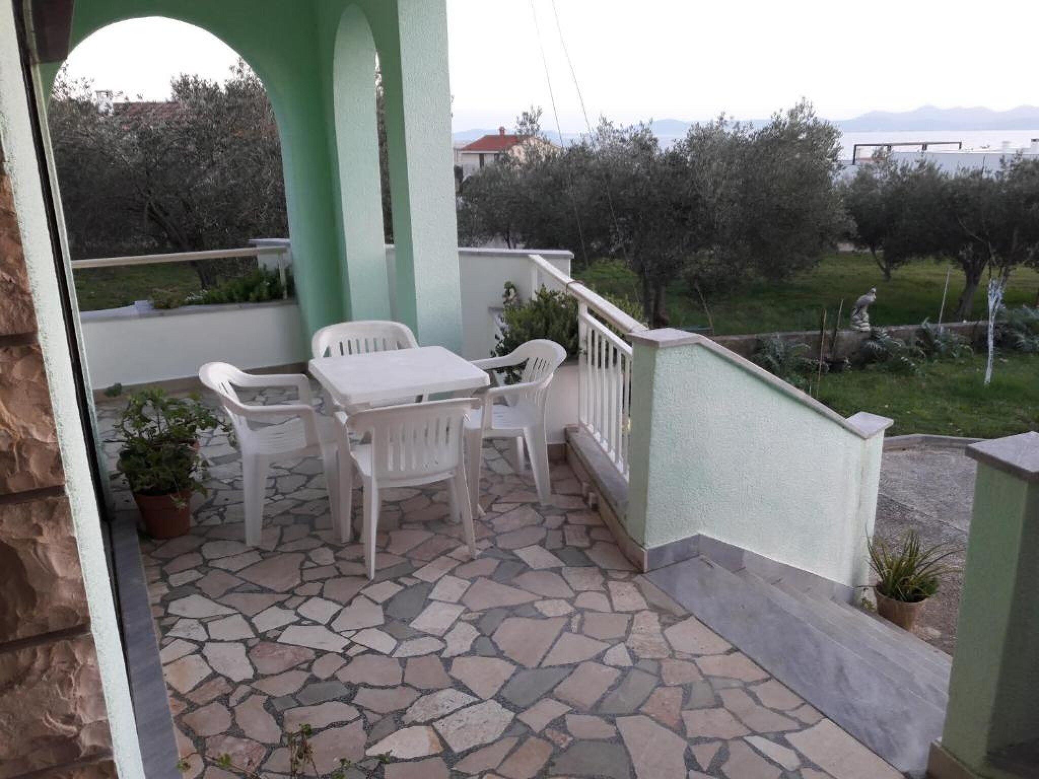 Eine Terrasse mit Steinboden, weißen Kunststoffstühlen und einem Tisch.
