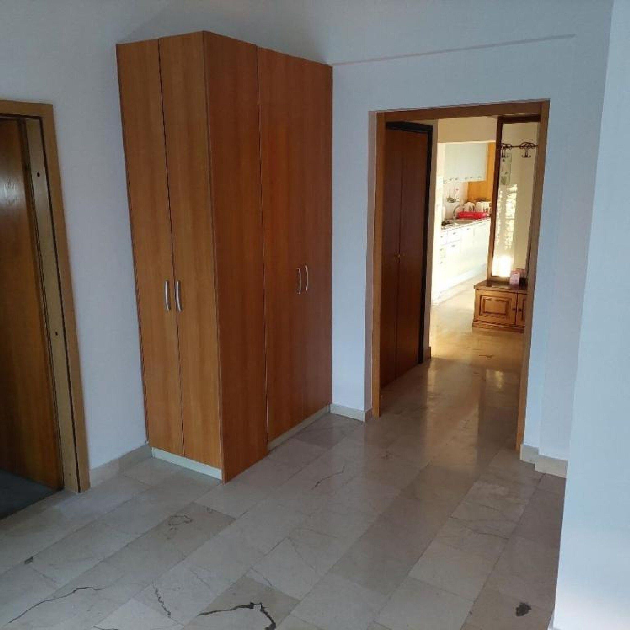 Une pièce dotée d’une grande armoire en bois, d’un sol carrelé et d’une porte-fenêtre donnant sur une autre pièce qui comprend un coin cuisine.