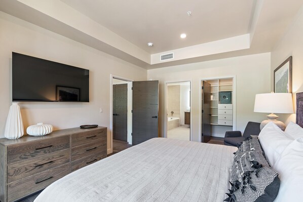 A modern bedroom with a large bed, a dresser, a TV mounted on the wall, and a walk-in closet.