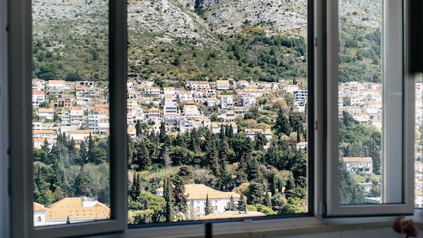 Un coin repas avec une table et des chaises, un vase rempli de fleurs et une vue sur une ville située sur une colline, visible à travers de grandes fenêtres.