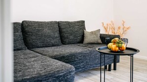 Un salon moderne avec un canapé d’angle gris, une petite table ronde sur laquelle est posé un panier de fruits et une plante décorative.
