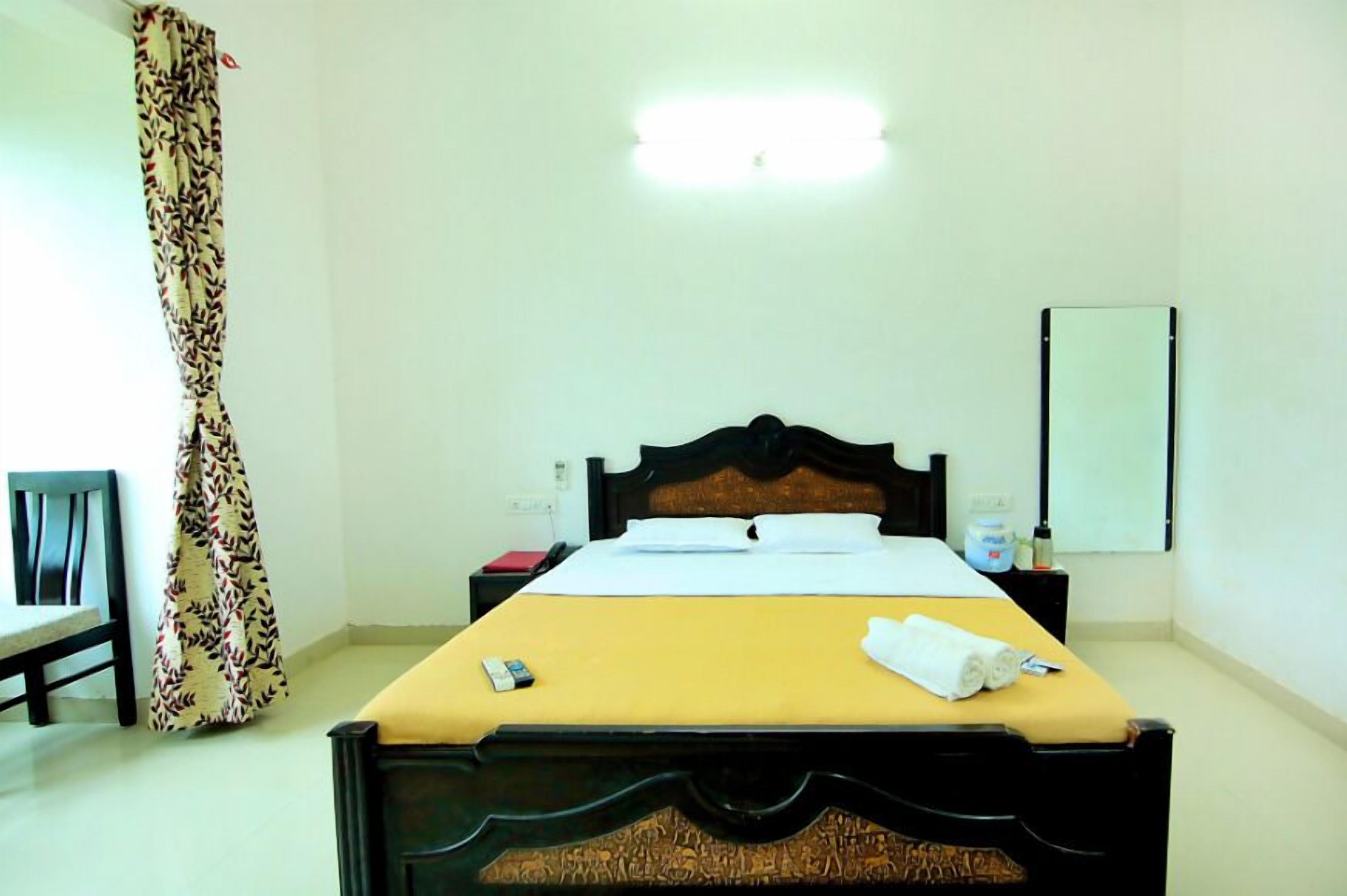 Habitación de hotel con cama, mesita de noche, lámpara, espejo, cortina y una silla.