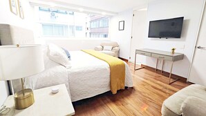 Une chambre d’hôtel équipée d’un lit, d’une table de chevet, d’une lampe, d’une télévision et d’une fenêtre donnant sur des immeubles.