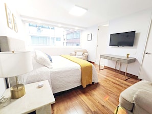 A hotel room with a bed, bedside table, lamp, TV, and a window with a view of buildings.
