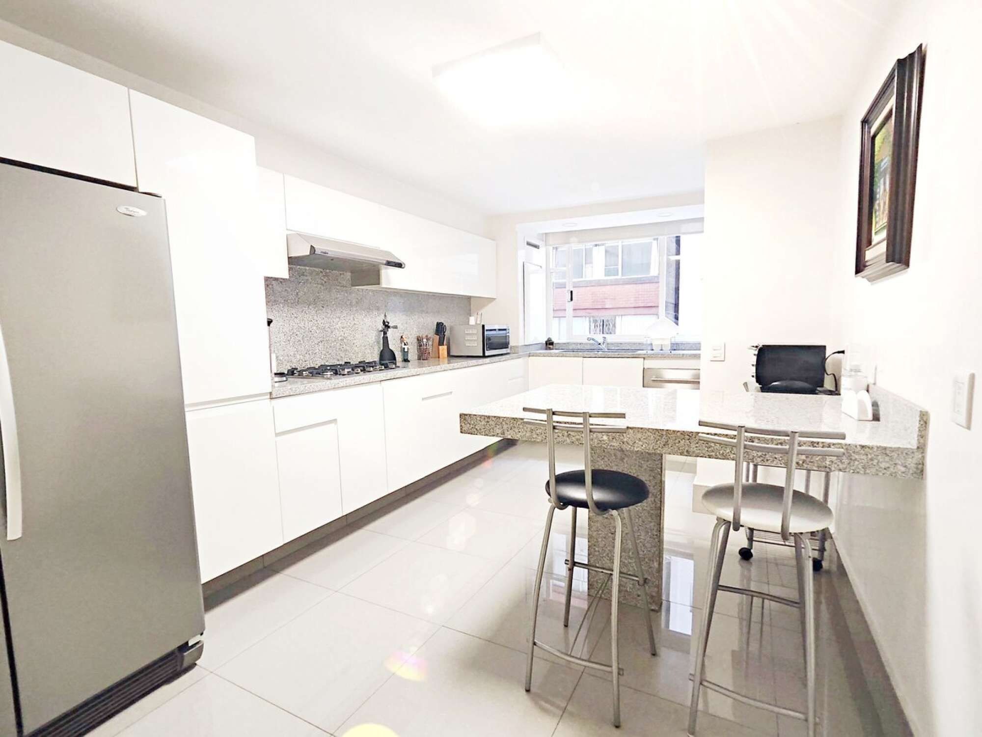 A modern kitchen with white cabinets, a stainless steel refrigerator, a granite countertop, and two bar stools.