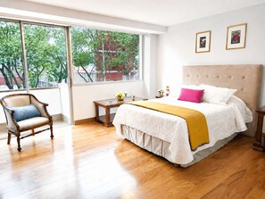 Ein Schlafzimmer mit großem Fenster, einem Bett mit weißer Bettwäsche und einer gelben Decke, einem Holzstuhl und einem kleinen Tisch mit Dekoration.