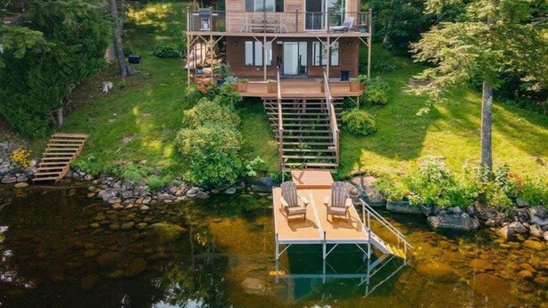 A wooden cabin with a deck and a dock extending into a lake, surrounded by trees. - Red Cedar Haven Lakefront Dock  Kayaks (Sidney)