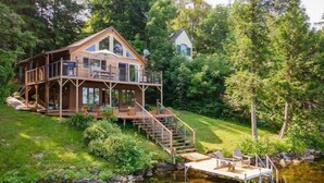 A wooden cabin with a deck and stairs leading to a dock over a pond, surrounded by lush greenery. - Red Cedar Haven Lakefront Dock  Kayaks (Sidney)