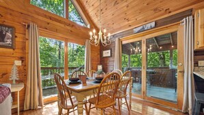 A wooden cabin with a dining area featuring a table and chairs, large windows, and a chandelier.