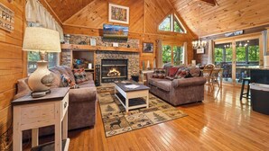 A cozy cabin interior with a stone fireplace, wooden furniture, and a large window overlooking greenery.
