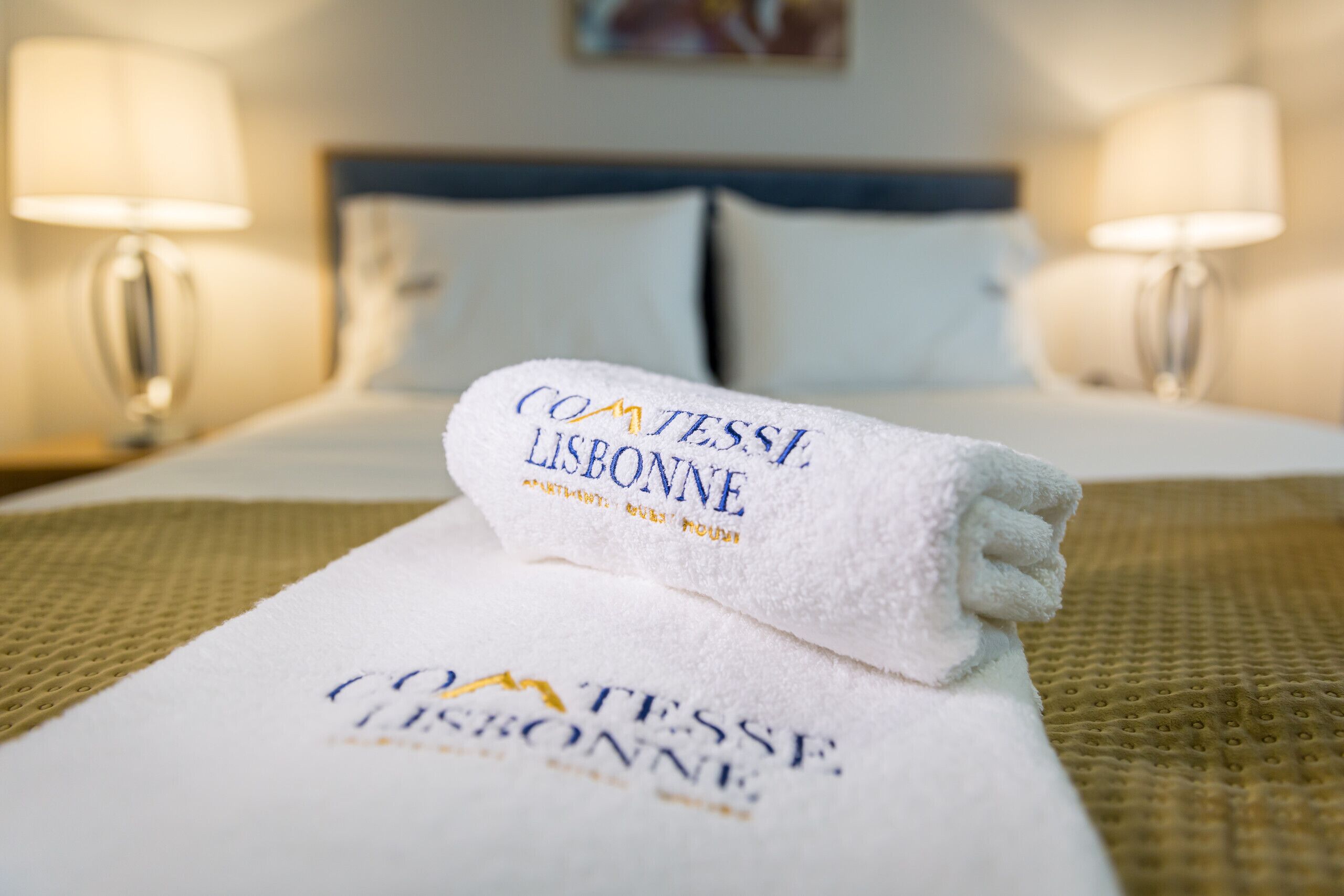 A neatly made bed with a folded towel on top, a headboard, and two bedside lamps.