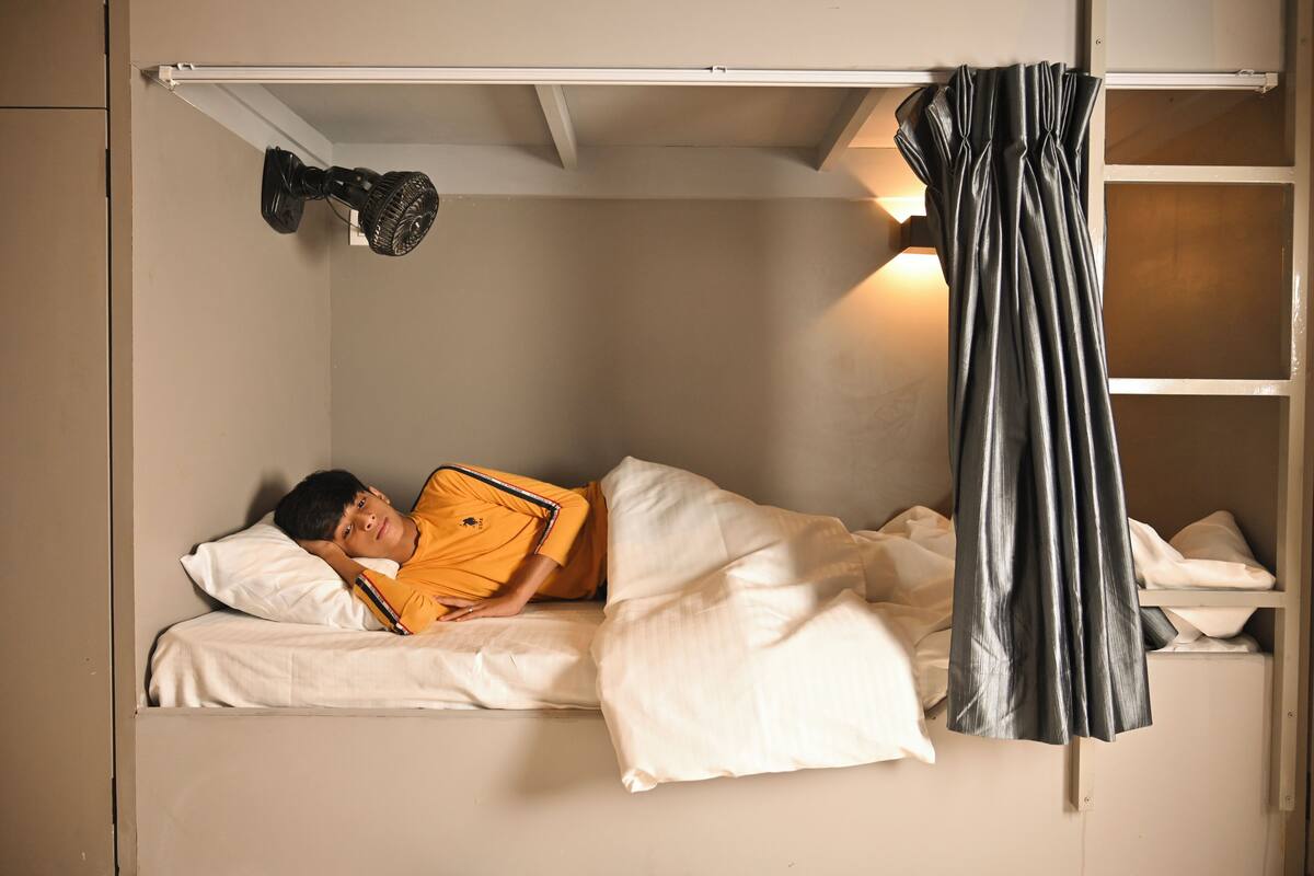 Una cama individual con ropa blanca, un ventilador de techo y una cortina negra.