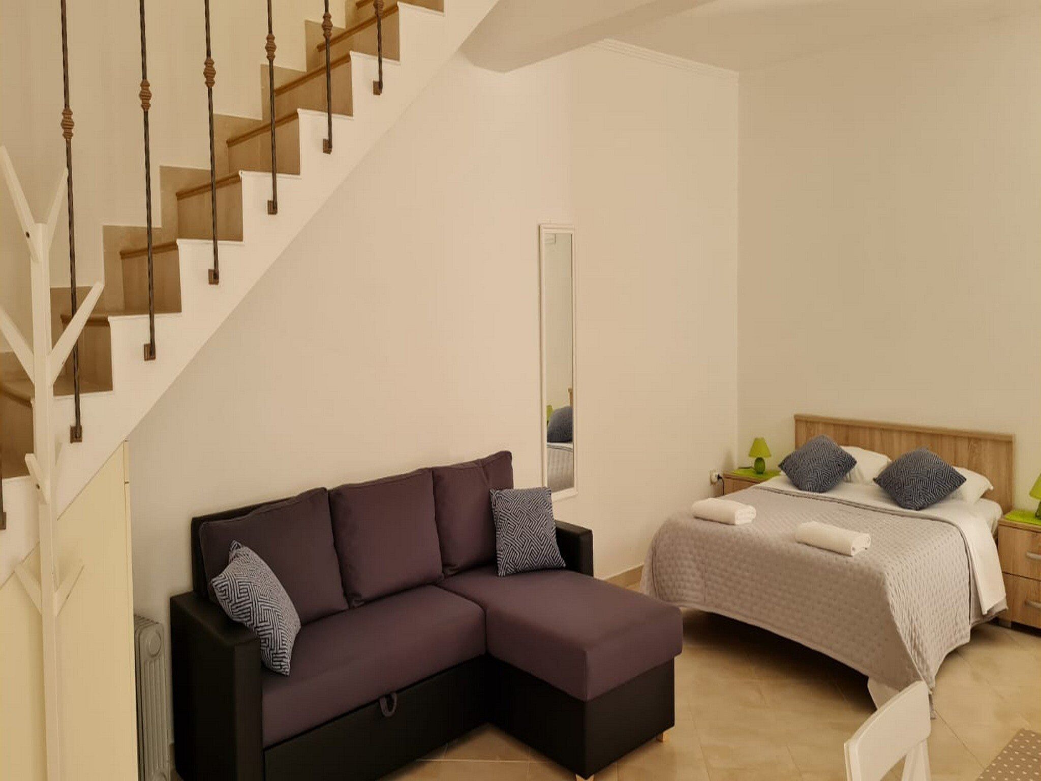 A modern bedroom with a sectional sofa, a bed with patterned pillows, a nightstand, and a staircase.