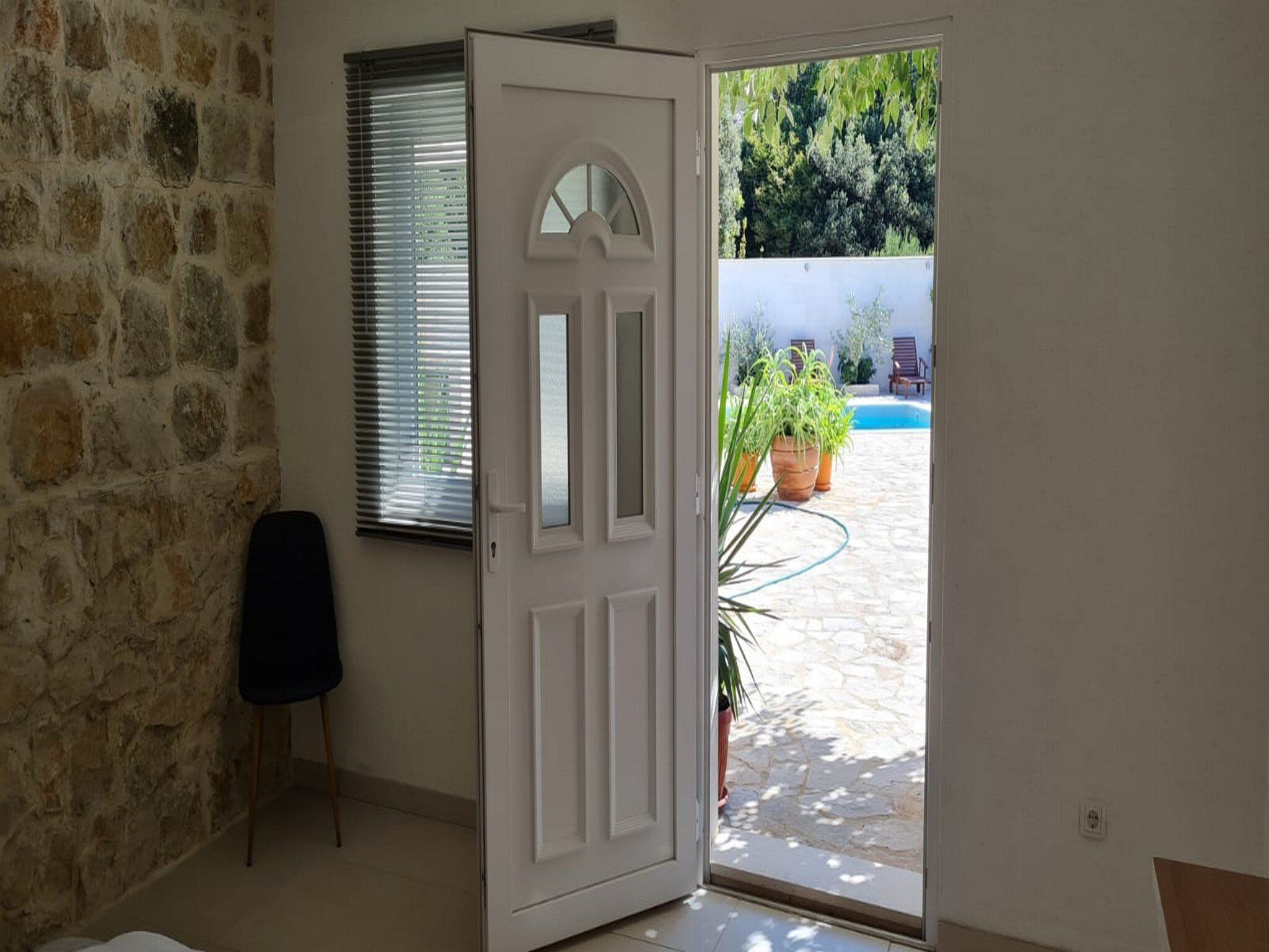 Habitación con pared de piedra, una puerta blanca, ventana con persianas y vista a una piscina y mobiliario exterior.