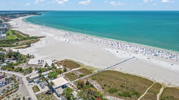 Vista aérea de una playa con una extensa franja de arena, algunos edificios y un cielo azul despejado.