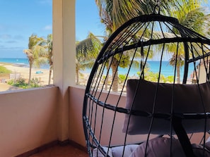 A wicker chair with a cushion, a balcony with a view of the beach and palm trees, and a clear blue sky.