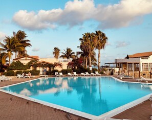 A pool area with lounge chairs, palm trees, and a building with a tiled roof.