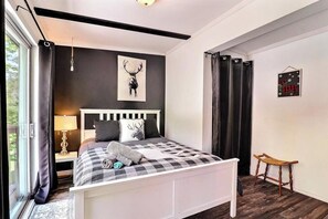 Quarto com uma cama grande, cabeceira, janela com cortinas, mesa de cabeceira com abajur e uma obra de arte na parede.