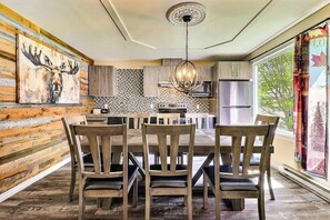 A dining area with a wooden table and chairs, a chandelier, and a kitchen in the background.