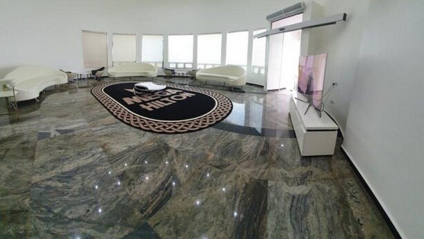 Un amplio y moderno vestíbulo de hotel con una alfombra circular que muestra el nombre del hotel, piso de mármol y un área de descanso con sofás blancos.