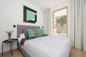 Ein Schlafzimmer mit einem Bett, einem Nachttisch mit Blumenvase, einem Fenster mit Vorhängen und einem Wandbild.