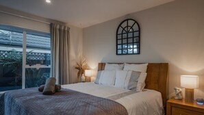 A bedroom with a large bed, a nightstand with a lamp, a window with curtains, and a wall-mounted mirror.