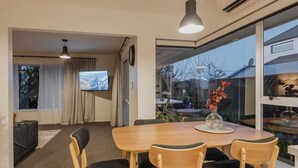 A dining area with a wooden table and chairs, a TV in the background, and a view through large windows.