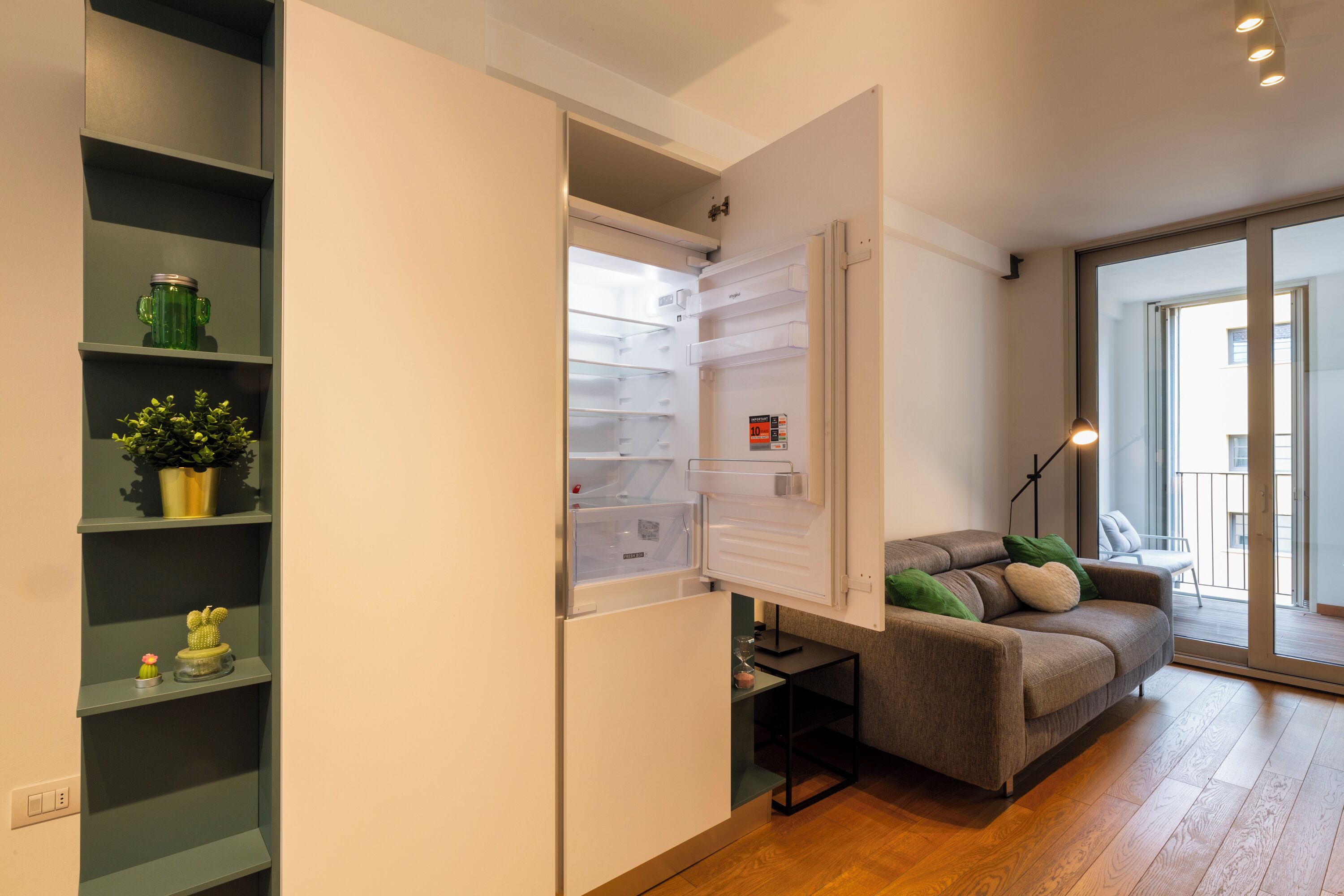 A modern kitchen with a built-in refrigerator, a wooden floor, and a grey sofa with cushions.